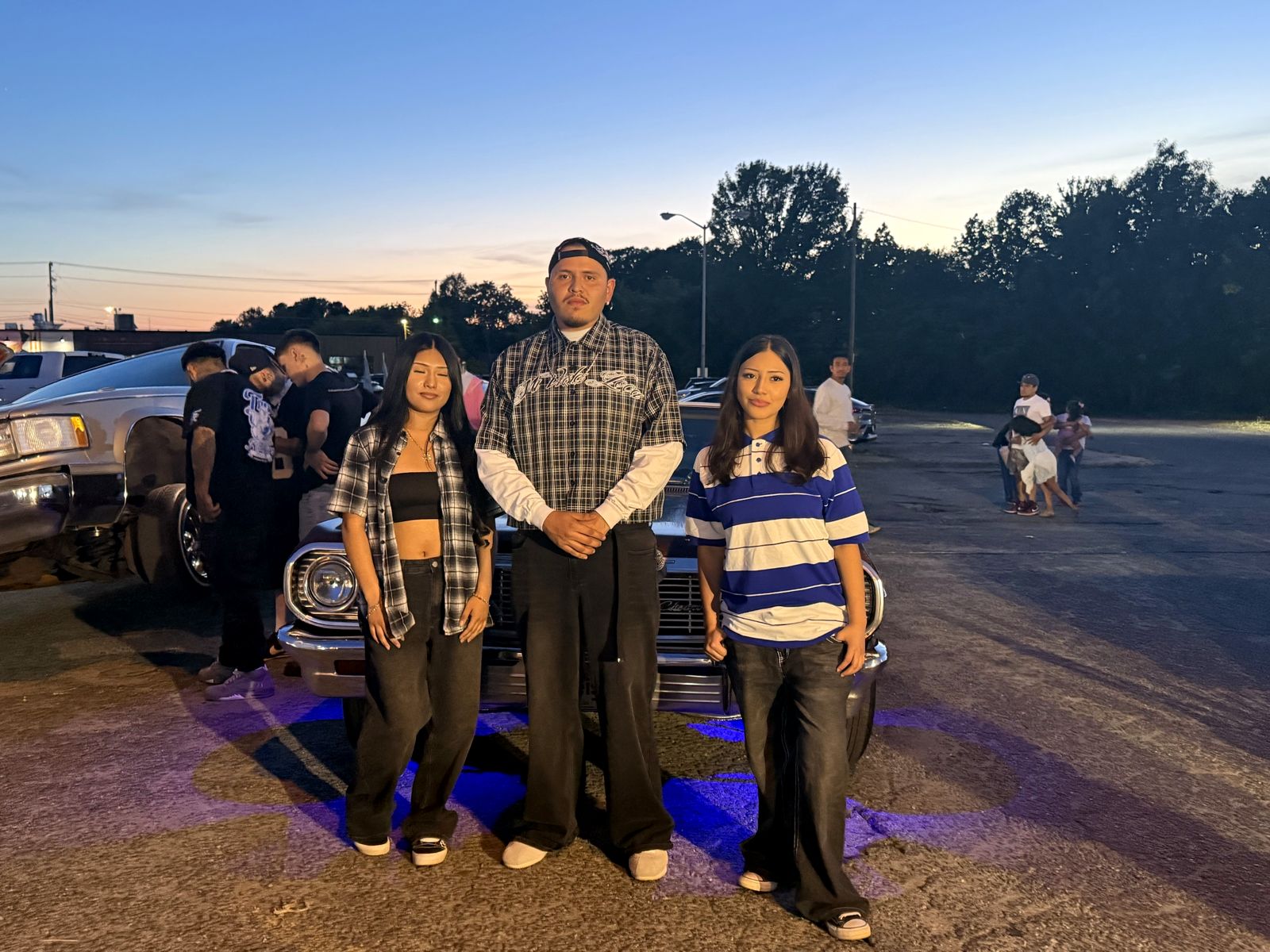 Three friends posing in front of a classic lowrider at dusk at Gatito Night Market