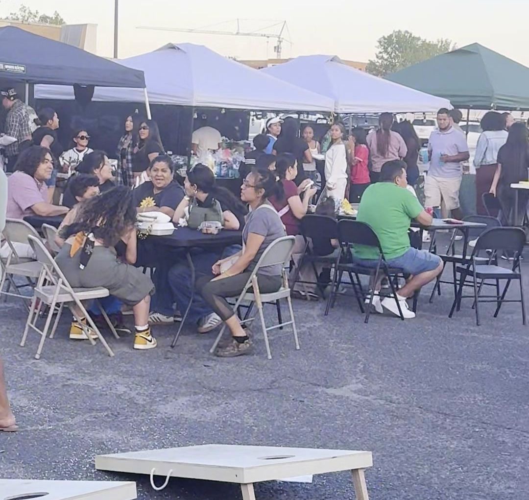 Families eating at tables under white and green vendor tents, with cornhole boards in front
